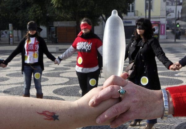 primer plano de un preservativo sujetado por dos manos y al fondeo varias mujeres de la mano con los ojos tapados