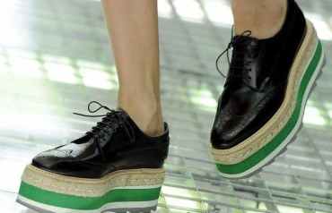 Primer plano de unos pies con zapatos negros con una gran cuña verde, blanca y esparto. Andan sobre una rejilla de metal.