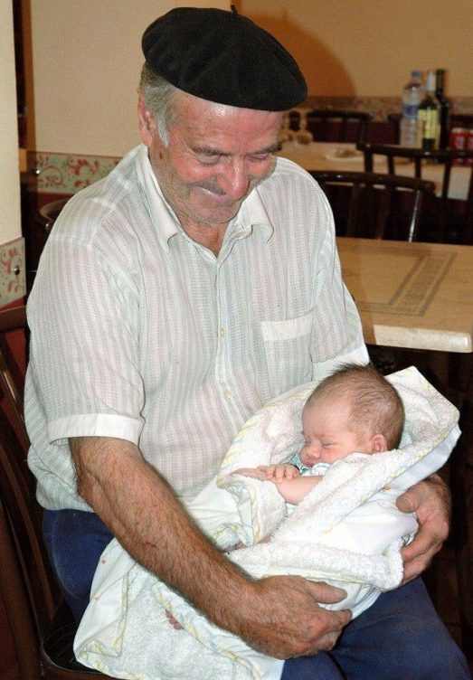 Un sonriente anciano, con boina, sujeta en sus brazos a un bebé, que duerme plácidamente.