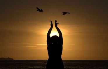 Una mujer alza sus manos hacia el sol naciente en una playa de Brasil a la vez que dos pájaros salen volando casi desde sus manos