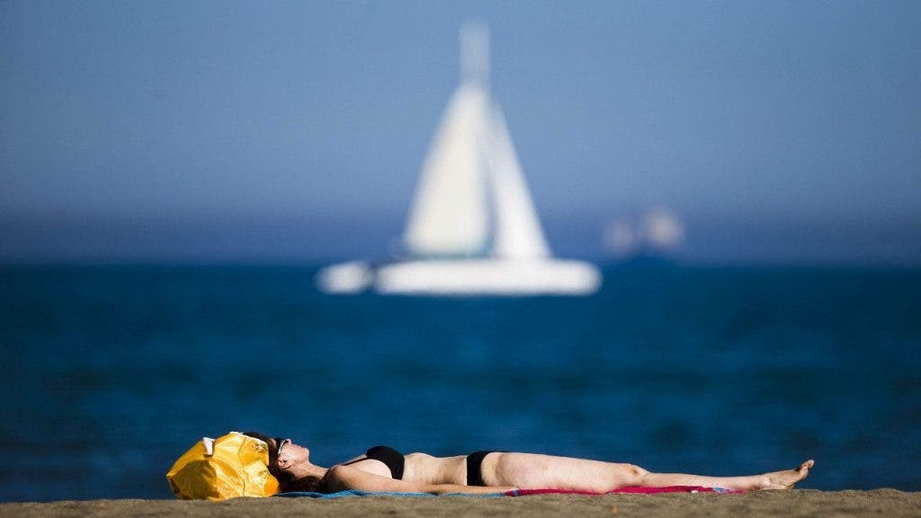 Una mujer toma el sol en biquini tumbada sobre su toalla en la playa de la Malagueta. Al fondo se ve un barco de vela que surca por el mar en calma. efesalud