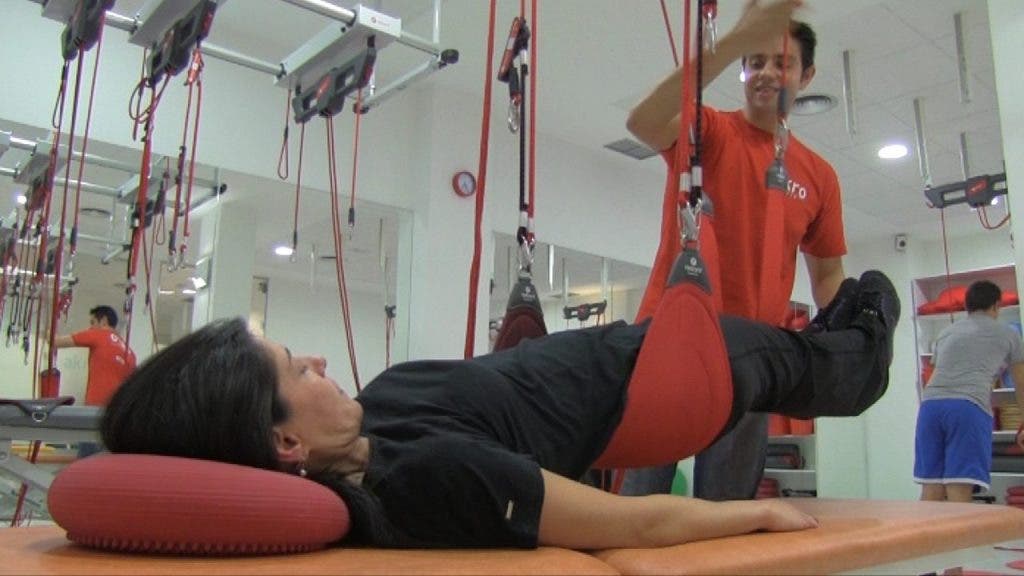 El fisioterapeuta trabaja con las cuerdas de Redcord la musculatura de Isabel, que está tumbada en la camilla con las dos piernas y la zona lumbar elevadas sobre la horizontal de la camilla