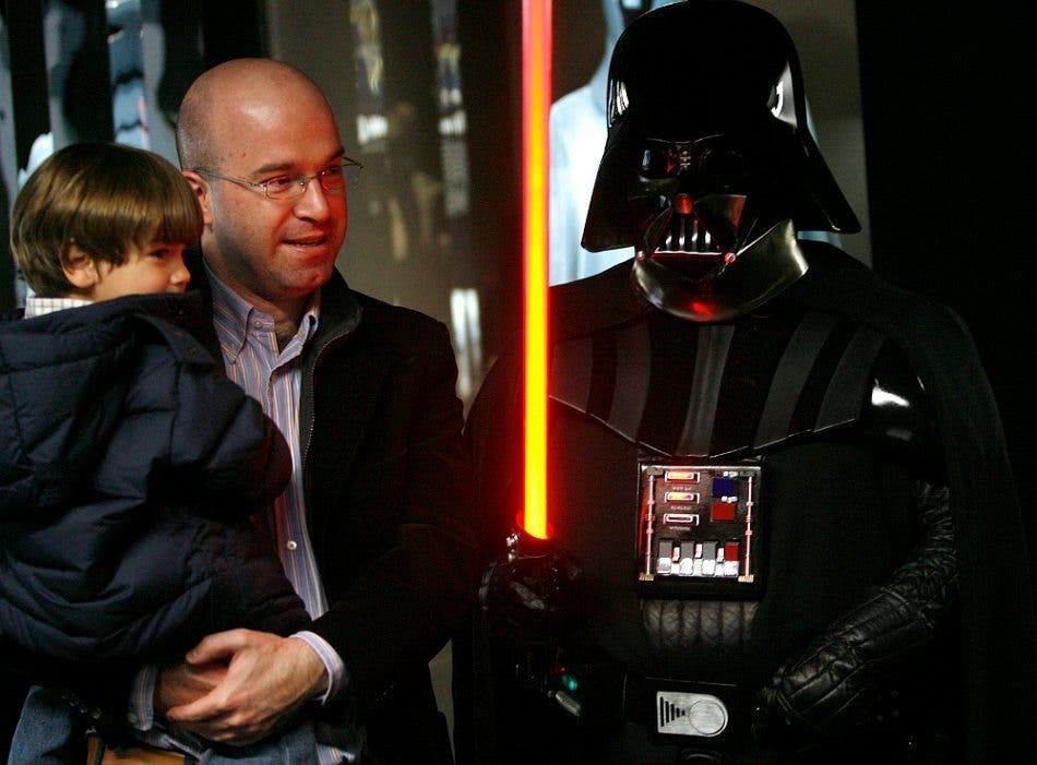 Un padre con su hijo en brazos contemplan la figura del malvado Darht Vader de la saga de La Guerra de las Galaxias, quien luce su mítica capa, casco y máscara negras y ase el no menos mítico rayo láser de color fuego