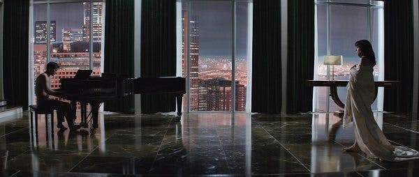 Escena de la película 50 Sombras de Grey donde aparece un hombre con el torso desnudo tocando el piano y una mujer se aproxima hacia él envuelta en una sábana, en el fondo de la imagen hay unos ventanales con vistas a rascacielos. Efesalud.com