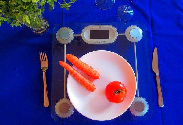 Composición donde, sobre un fondo azul eléctrico, se ve un peso a modo de bajoplato con un plato blanco encima que contiene dos zanahorias y un tomate. A los lados, un cuchillo a la derecha y un tenedor a la izquierda.